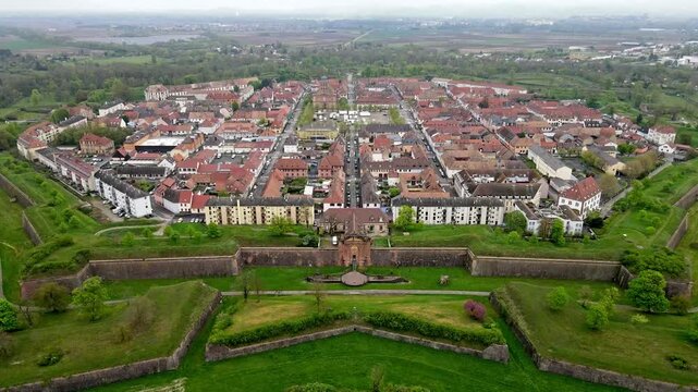 Aerial drone view of octagonal star fortress with pointed bastions and green moat under overcast sky. Terracotta rooftops and grid streets within fortification walls with Alsace countryside beyond.