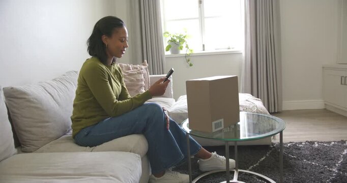 African American woman tapping smartphone on sofa after notification, glancing at sealed parcel