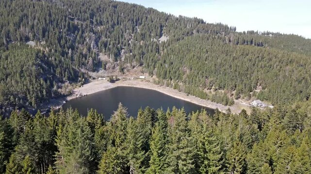 Aerial drone view of Lac Noir mountain lake ringed by dense conifer forest with a small dam and buildings on the far shore in the Vosges Alsace. Scenic alpine lake and nature travel.