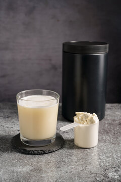 Protein vanilla shake in drinking glass and scoop with whey protein powder on dark background