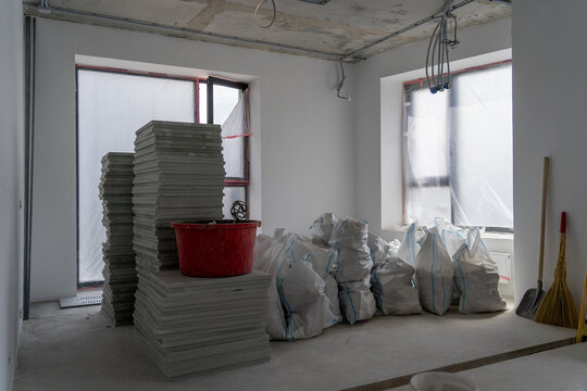 Modern apartment interior under renovation with stacks of ceramic floor tiles, construction debris bags, red mixing bucket, and tools in raw unfinished room ready for finishing work.