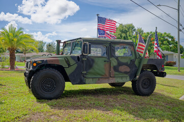 Fototapeta premium Camouflaged military Humvee (HMMWV) parked on a suburban lawn with several American flags and banners. Editorial Use Only July 5, 2026 St. Petersburg , FL USA. Evoking patriotism, veterans, and milita