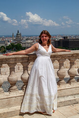 Fototapeta premium Smiling young woman in white summer dress poses on stone balcony overlooking historic cityscape on bright sunny day during her beautiful European summer vacation travels