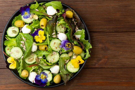 Colorful salad with greens cucumbers olives cheese and pansies flowers on a wooden table