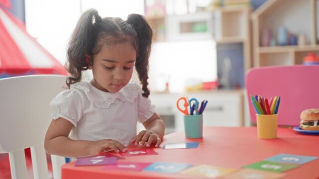 Girl placing number cards with right hand at red table in preschool building; concentration early learning.