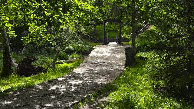 Lush green summer forest with a narrow stone path leading to a small suspension bridge over a tranquil stream, bathed in dappled sunlight filtering through the vibrant tree leaves