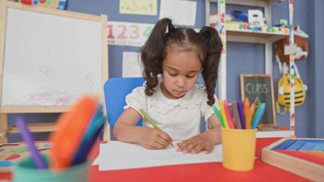 Little girl drawing with green pencil, right hand on paper at red activity table in a school building classroom filled with art supplies; concentration creativity.
