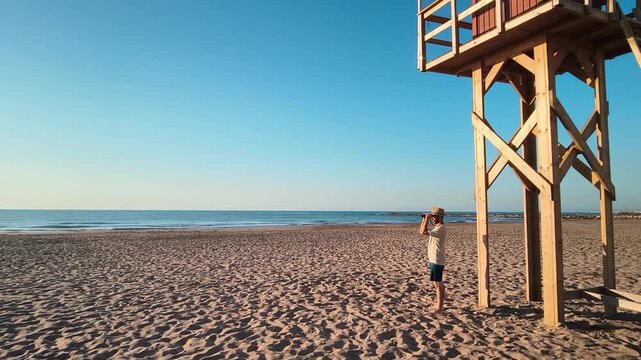 A man wearing a hat stands on the sandy beach using binoculars to observe the sea, with the wooden structure of a lifeguard tower visible behind him, representing coastal surveillance and ansafety