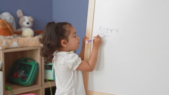 Little girl draws a sun and writes letters on a whiteboard while holding a purple marker in a playroom; concentration creativity play.