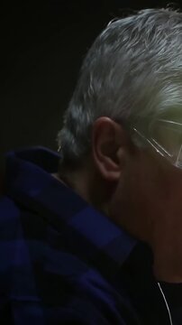 Vertical video: Tilting man shifting jaw as light changing in dim room, glasses earbud plaid collar