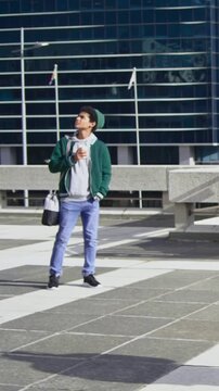 Vertical video: Walking man starting, scanning, carrying bag and coffee in plaza with green hoodie
