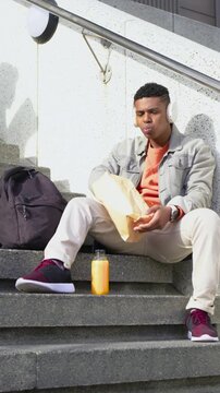 Vertical video: Sitting man with headphones unpacking lunch and sipping juice during break on steps