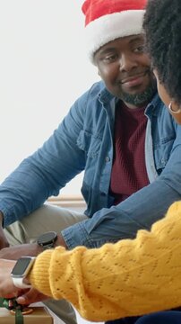 Vertical video: Watching woman in yellow knit undoing bow, man in Santa hat steadying box on couch
