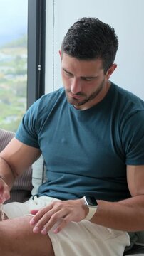 Vertical video: Using dropper, man in teal T rubbing serum into hands to hydrate by window watch
