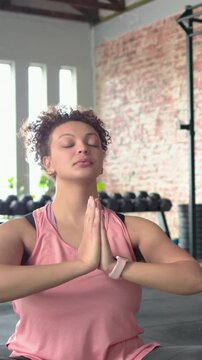 Vertical video: Inhaling, raising arms, woman in pink top practicing yoga at gym, dumbbells visible
