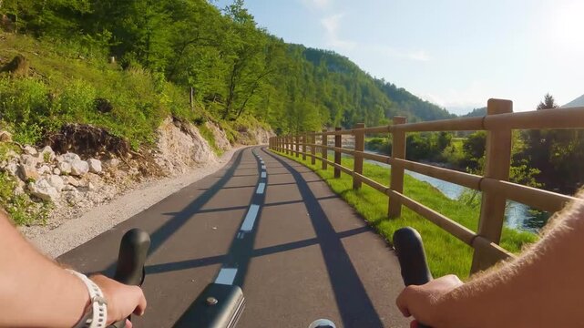 POV of cyclist riding on asphalt bike path at sunset in summer. Slovenia. First person view of male hands on bicycle handlebar cycling on road, wooden fence, river, green forest. Active sport outdoor