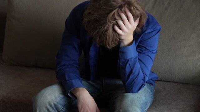Young man (zoomer) with messy hair leaning forward
 and holding his head. Visibly troubled, feeling stress, burnout and mental exhaustion. Depression and mental health concept.