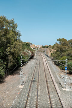 Twin electrified railway tracks curve through a sunlit Mediterranean landscape surrounded by mature trees and distant hillside homes beneath an expansive clear blue sky. Strong leading lines and overh