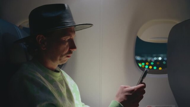 Airplane cabin interior with fashionable passenger checking mobile phone as aircraft approaches landing. Bokeh city lights visible through window at night while traveler browses device in seat.