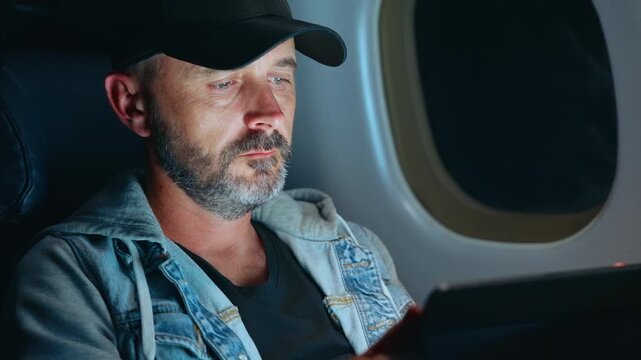 Middle-aged man with gray beard wearing black cap and denim jacket uses digital tablet while seated in airplane cabin. Screen glow illuminates his focused face near window during flight journey.