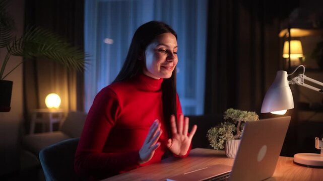 A young brunette woman in a red turtleneck sits at a wooden desk at night, smiling and gesturing with open hands during a video call or online meeting via her laptop in a cozy home office