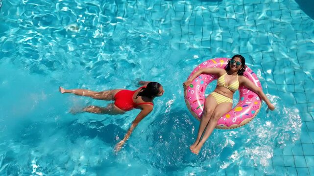 Two young women enjoying summer vacation in swimming pool, one floating on donut ring and one swimming, aerial view