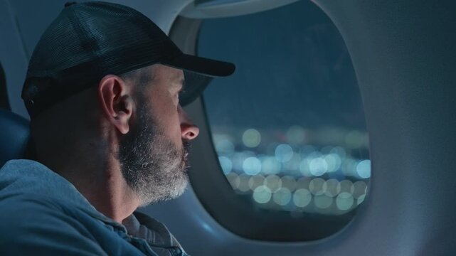 Airplane cabin interior showing middle-aged bearded man wearing baseball cap looking through window as plane approaches landing. Bokeh city lights visible outside during night flight arrival.