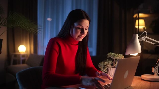 A tired young brunette woman in a red turtleneck touches her forehead, looking exhausted while working on her laptop late at night in a cozy home office, suffering from a headache.