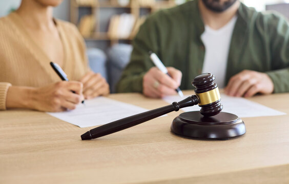Couple sign divorce document beside court gavel. Hands add a signature to the agreement as a lawyer guides paperwork and resolves breakup conflict. Concept shows legal separation and justice.