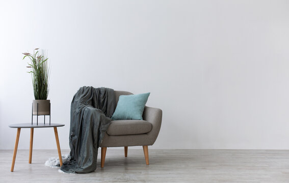 Modern boho cozy corner of home in living room. Gray vintage armchair with blue pillow and blanket, table with green plant in pot, wooden floor, on light wall background, copy space, mockup, nobody