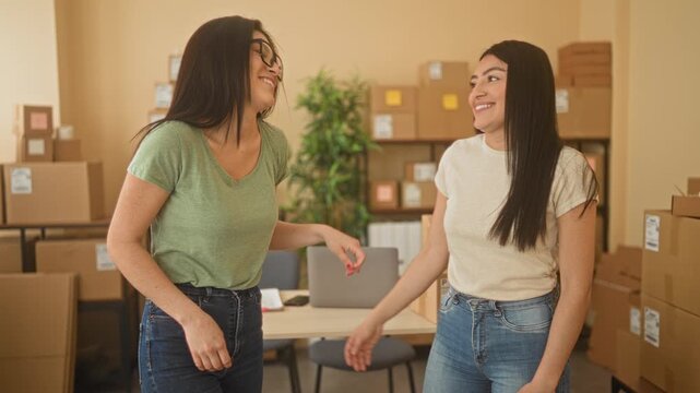 Two women hug with arm around shoulder, smiling beside stacked cardboard boxes and parcels at a small business building; friendship entrepreneurship joy.