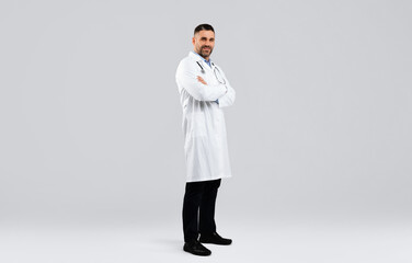 Cheerful middle aged man doctor posing with folded arms looking at camera standing on light...