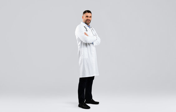 Cheerful middle aged man doctor posing with folded arms looking at camera standing on light background, wearing white uniform, coat and stethoscope, full body length, free space