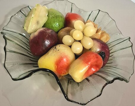 Fruteira de vidro ondulado com arranjo decorativo de frutas artificiais coloridas sobre mesa clara, destacando uvas, caju e goiaba em composi&ccedil;&atilde;o de natureza-morta para decora&ccedil;&atilde;o de interiores.