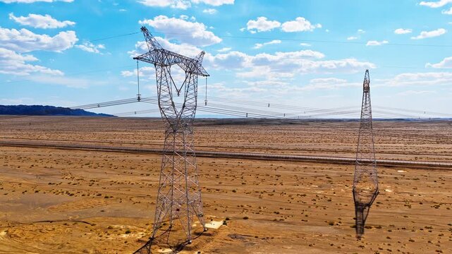 High-voltage power transmission towers standing in a vast desert landscape under a blue sky with clouds.