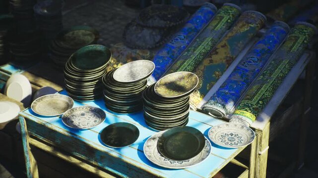 Sunlit souk stall with ceramic plates in morocco market, colorful glazed plates and leaning tile rolls on rustic table, bold blue patterns, strong shadows