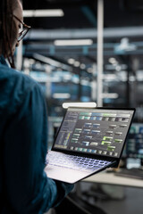Naklejka premium Data center administrator uses a laptop to investigate neural network configurations, working with AI technology and high performance systems.