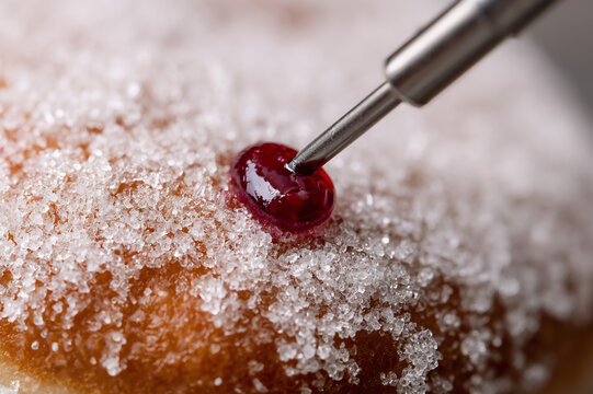 Macro photograph of a metal nozzle injecting vibrant raspberry jelly into a sugar-dusted donut