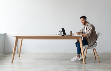 Profile portrait of young smiling Arab man in wireless headset using pc sitting at desk, writing in notebook. Cheerful guy browsing internet, watching webinar studying online, free copy space © Prostock-studio