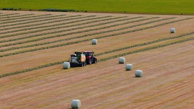 Aerial view of southern Iceland farmland near Skogafoss. A red tractor tows a baler, forming and wrapping silage bales on windrowed stubble under soft natural light.