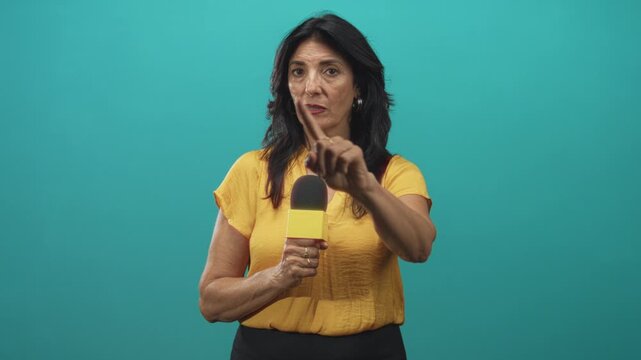 Woman reporter holding microphone points finger forward in studio wearing yellow blouse; assertive authority.