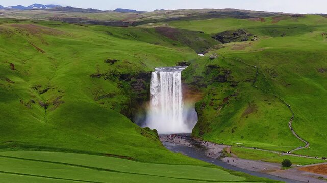 Aerial view shows Skogafoss in southern Iceland as a single drop falls between moss covered cliffs. A rainbow arcs in mist while visitors walk paths and riverbank.
