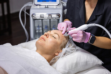 Fototapeta premium Cosmetologist performs a professional HydraFacial treatment using a hydradermabrasion machine to cleanse, exfoliate and hydrate facial skin in a modern beauty clinic.