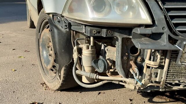 Tight view of a crushed silver car left-front corner parked at the curb, showing a broken bumper, bent hood, detached headlight and exposed radiator amid
