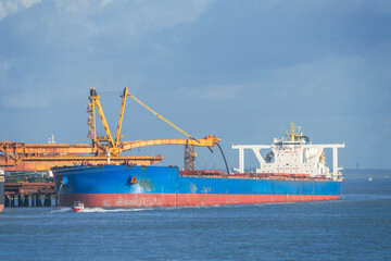 Fototapeta premium Blue bulk carrier is being loaded at an industrial port, with a yellow crane feeding material into its hold. A small pilot boat speeds nearby on calm waters