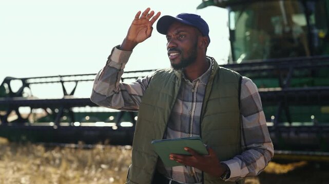 A man stands in a field near a large farming machine. He looks at a tablet to check information about crops. The sun shines on the farm in the background.
