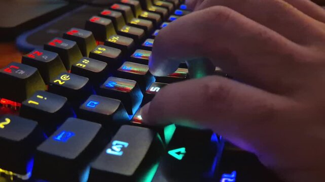 A close-up of a hand typing and gaming on a black, RGB backlit mechanical keyboard in a low light room is shown.