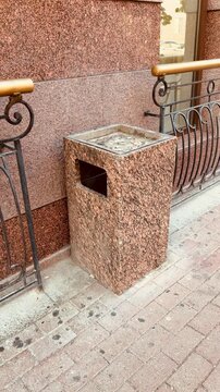 The image features a brown granite trash can with a metal slot on its side, positioned on a brick sidewalk. The can has a square top and is situated next to a black metal railing with wooden handrails
