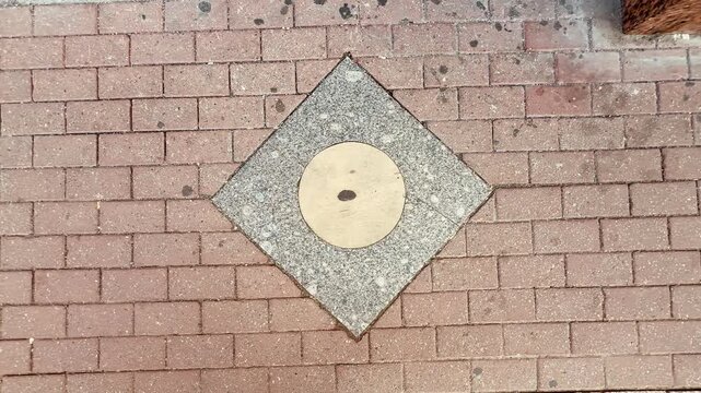Aerial view of a sidewalk plinth in a square. The plinth is circular with a central hole, surrounded by a diamond-shaped border of brick pavement