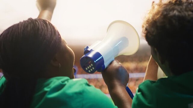 Football supporters chant with megaphone in packed sports arena, reacting to goal, showing woman shouting towards soccer pitch, creating loud stadium atmosphere for fan culture content.